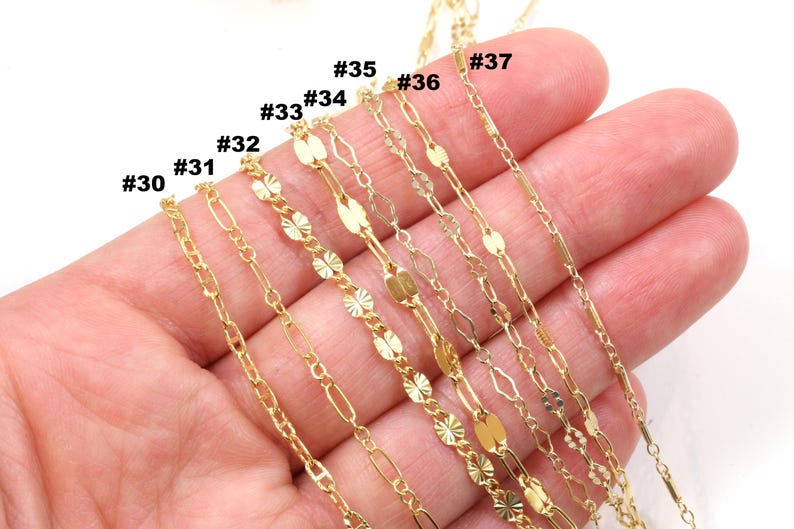 Peut inclure: Une s&eacute;lection de colliers cha&icirc;ne dor&eacute;s d&eacute;licats, chacun avec un design unique. Les cha&icirc;nes sont tenues dans une main, pr&eacute;sentant la vari&eacute;t&eacute; des styles, y compris les maillons ovales, les accents floraux et les formes allong&eacute;es. Les num&eacute;ros #30 &agrave; #37 sont visibles.