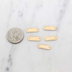 May include: Five gold-colored metal rectangular tags with two holes each. The tags have a textured design around the edges.