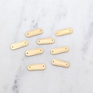 May include: Eight gold-toned metal rectangular tags with rounded corners and two holes. The tags have a textured surface.
