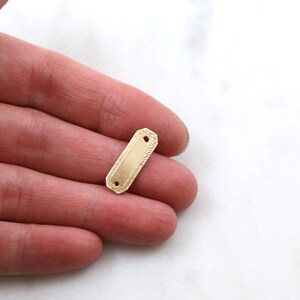 May include: A gold-colored metal tag with a rectangular shape and a textured surface. The tag has two holes for attaching it to something.