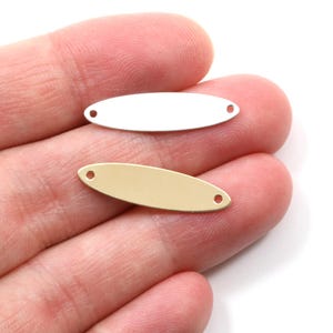 May include: Two oval-shaped metal jewelry tags. One tag is silver-colored, and the other is gold-colored. Each tag has two small holes, one on each end. The tags are displayed on a person's hand.