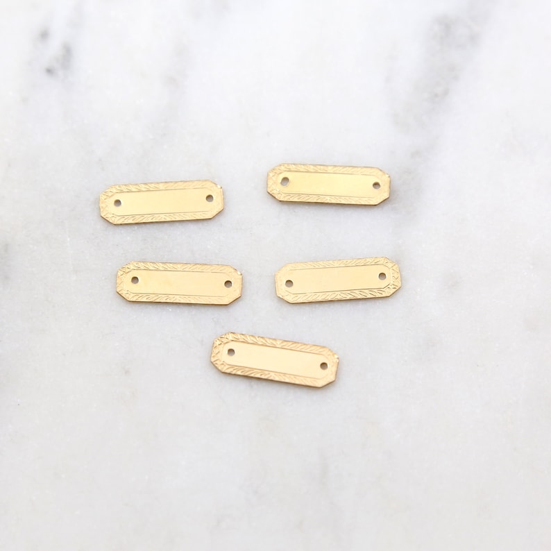 May include: Five gold-colored metal rectangular tags with two holes each. The tags have a textured surface and a slightly raised edge.