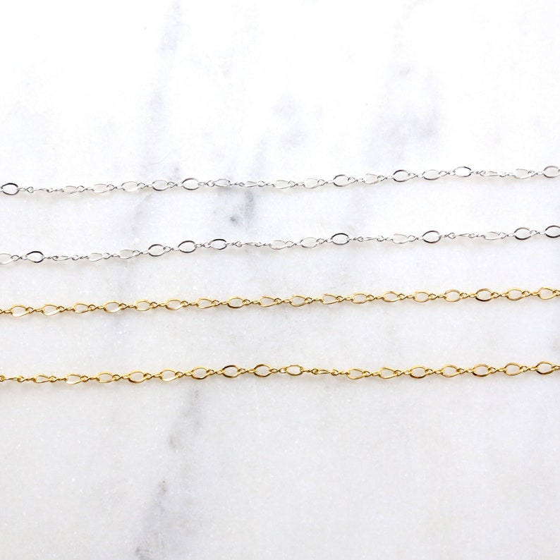 May include: Four chains of different lengths, two silver and two gold, are laid out on a white marble surface. The chains are all made of small, oval-shaped links.