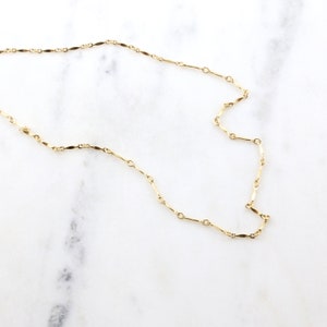May include: A delicate gold chain necklace with a simple, elegant design. The chain is made of small, flat links that are connected together in a continuous pattern.