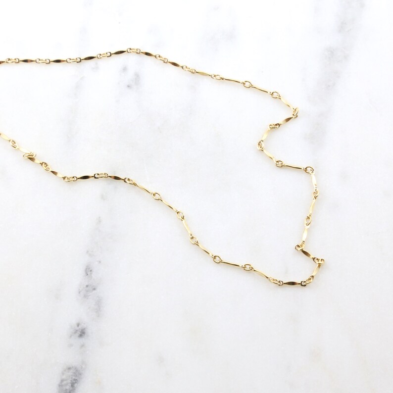 May include: A delicate gold chain necklace with a simple, elegant design. The chain is made of small, flat links that are connected together in a continuous pattern.