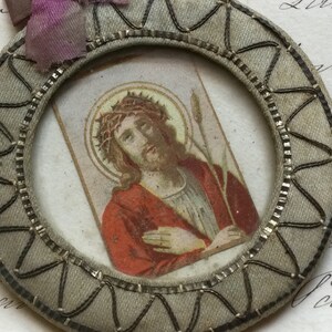 May include: A round, gray fabric frame with a silver trim and a small image of Jesus Christ inside. The image shows Jesus with a crown of thorns and a reed in his hand.