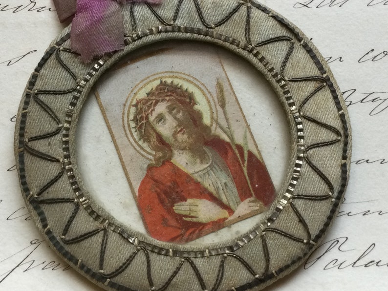 May include: A round, gray fabric frame with a silver trim and a small image of Jesus Christ inside. The image shows Jesus with a crown of thorns and a reed in his hand.
