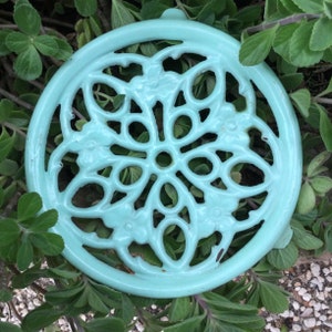 Puede incluir: Un posavasos redondo de hierro fundido de color azul claro con un diseño floral decorativo. El posavasos es redondo y tiene un borde elevado.