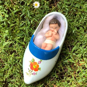 May include: A white wooden shoe with blue trim and floral designs, containing a small porcelain baby doll figurine lying on a white cotton bed.