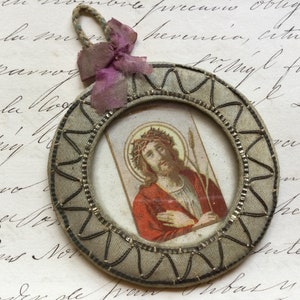 May include: A small, round, framed picture of Jesus Christ with a crown of thorns and a spear. The frame is made of fabric and has a decorative border. The picture is attached to a pink ribbon.