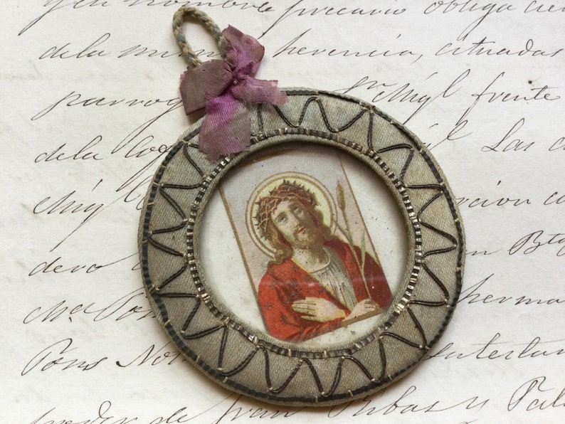 May include: A small, round, framed picture of Jesus Christ with a crown of thorns and a spear. The frame is made of fabric and has a decorative border. The picture is attached to a pink ribbon.