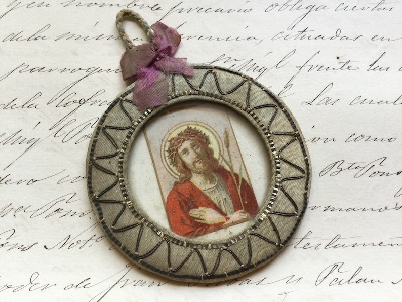 May include: A round, fabric-covered frame with a silver trim and a small image of Jesus Christ inside. The frame is decorated with a purple ribbon.