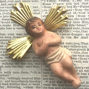 Puede incluir: Una figurita vintage de un ángel bebé con un halo de rayos dorados. El ángel está hecho de yeso y pintado de color marrón claro. El ángel lleva una tela blanca y tiene una dulce expresión en su rostro. La figurita está colocada sobre una página de un libro con texto.