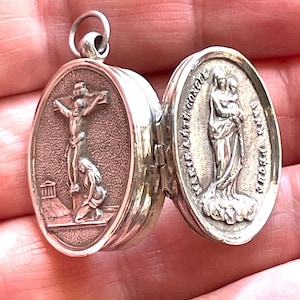 Pode incluir: Um pingente de medalhão oval de prata com imagens religiosas. Um lado retrata a crucificação de Jesus, com uma figura ajoelhada abaixo. O outro lado mostra uma mulher segurando uma criança. O medalhão está aberto.