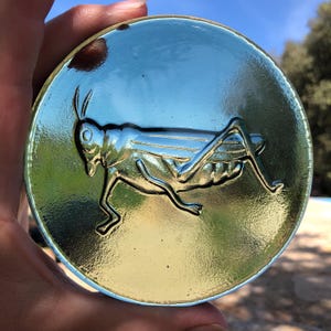 May include: A round, glass trinket with a silver-coloured grasshopper design. The glass is a light blue-green colour.