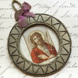 May include: A round, framed image of Jesus Christ wearing a crown of thorns and holding a reed. The frame is made of fabric and has a decorative border. The image is attached to a pink ribbon.