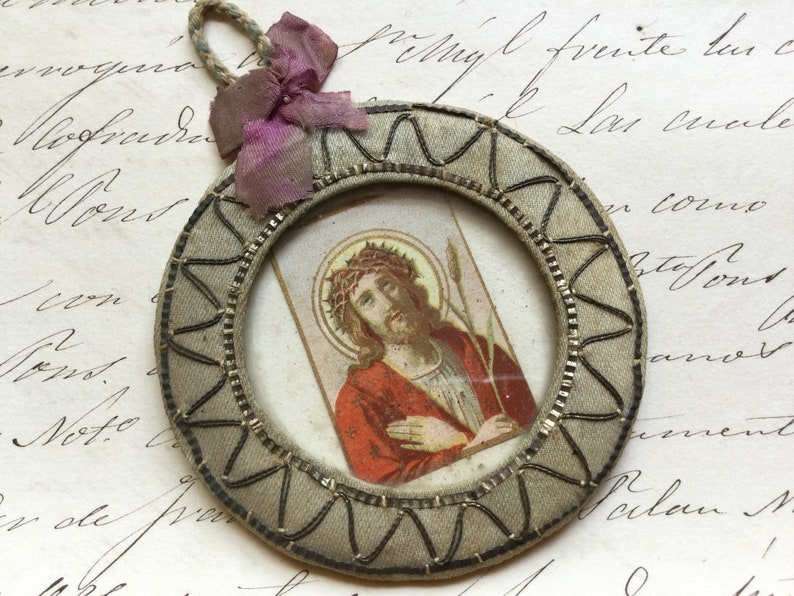 May include: A round, framed image of Jesus Christ wearing a crown of thorns and holding a reed. The frame is made of fabric and has a decorative border. The image is attached to a pink ribbon.
