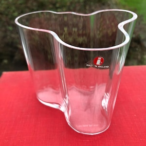 May include: A clear glass vase with a wavy, organic shape. The vase has a red sticker on the side that says "Made in Finland".