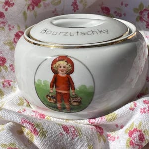 May include: A white ceramic jar with a lid, featuring a gold rim and the word "Bourzutschky" in gray lettering. The jar has a circular illustration of a child in red clothing carrying baskets. The jar is sitting on a floral patterned cloth.