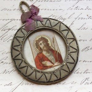 May include: A small, round, framed image of Jesus Christ wearing a crown of thorns. The frame is made of fabric and has a silver and black design. The image is on a white background.