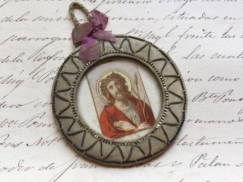 May include: A small, round, framed image of Jesus Christ wearing a crown of thorns. The frame is made of fabric and has a silver and black design. The image is on a white background.
