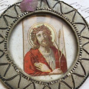 May include: A round, gray fabric medallion with a silver-colored metal frame. The frame encloses a small, rectangular image of Jesus Christ wearing a crown of thorns and holding a reed. The image is on a white background with a gold halo around Jesus' head.