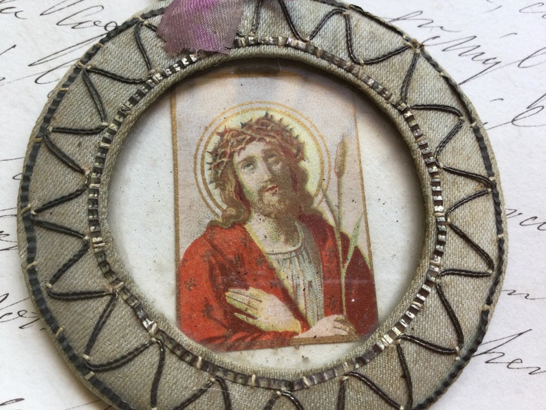 May include: A round, gray fabric medallion with a silver-colored metal frame. The frame encloses a small, rectangular image of Jesus Christ wearing a crown of thorns and holding a reed. The image is on a white background with a gold halo around Jesus' head.