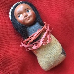 May include: A handmade doll with a brown face, black hair, and a white headband. The doll is wrapped in a brown and white fabric and tied with pink ribbon.