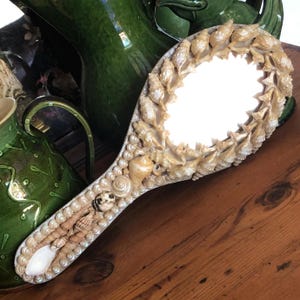 Puede incluir: Un espejo de mano con un mango plateado y un espejo redondo rodeado de conchas marinas. Las conchas marinas son de varios colores, incluyendo blanco, marrón y beige.