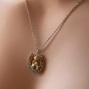 Puede incluir: Collar dorado con un colgante de herradura con una pequeña figura en su interior. La herradura está adornada con piedras preciosas de colores. El collar lo lleva una persona.