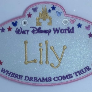 Op de afbeelding: Een wit ovaal naambadge met een roze rand, op een gouden glitterachtergrond. Het badge heeft een paarse geborduurde "Walt Disney World" tekst, een gouden geborduurd kasteel en een gouden geborduurde naam "Lily". De onderkant van het badge heeft "Where Dreams Come True" in paarse borduurwerk. Het badge is versierd met paarse, roze en blauwe sterren en Mickey Mouse-koppen.