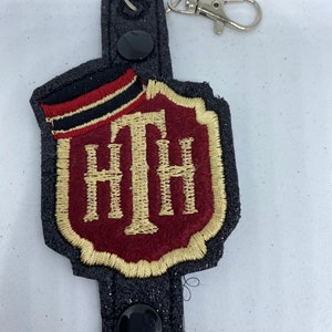 Op de afbeelding: Een zwarte en gouden geborduurde sleutelhanger met een rood en gouden geborduurd logo met de tekst "HTH". De sleutelhanger heeft een zwarte stoffen band met twee zwarte knopen.