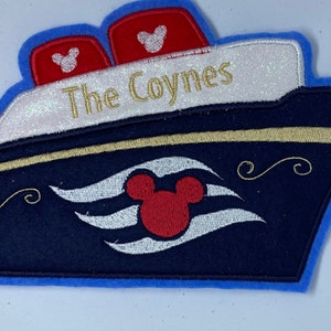 May include: A blue and white embroidered patch shaped like a cruise ship with the text "The Coynes" on the top. The ship has a Mickey Mouse head with red ears and white eyes on the front. The ship has a white border and a gold trim.