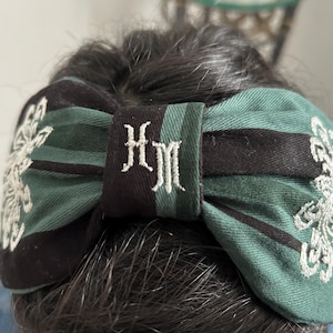 Puede incluir: Una diadema a rayas verdes y negras con un lazo bordado en blanco y las iniciales "HM" en el centro. La diadema tiene un diseño bordado en blanco a cada lado.