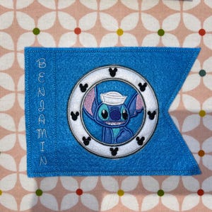 May include: Blue felt pennant with the name "Benjamin" embroidered on the left side. A white circle with a blue Stitch character wearing a sailor hat is in the center.