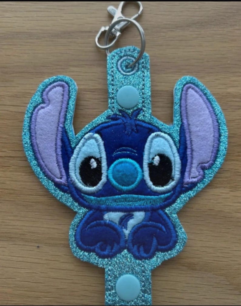K&ouml;nnte beinhalten: Ein blauer und violetter Stitch-Schl&uuml;sselanh&auml;nger mit einem silbernen Verschluss. Der Schl&uuml;sselanh&auml;nger hat eine glitzernde blaue Kontur und ein detailliertes gesticktes Design der Figur Stitch aus dem Disney-Film Lilo & Stitch. Der Schl&uuml;sselanh&auml;nger hat einen Knopfverschluss.