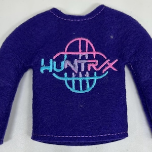 May include: A dark purple long-sleeved top featuring the embroidered word "HUNTRAX" in a colourful design. The design incorporates pink, blue, and teal hues. The top has a crew neck and is made of a soft material.