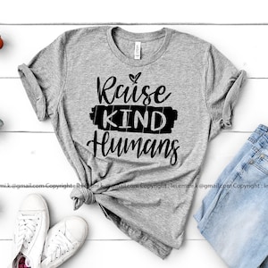 May include: A heather gray t-shirt with the phrase "Raise KIND Humans" in black script. The shirt is styled with light blue jeans and white sneakers. Small potted succulents are arranged around the clothing.