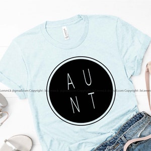 May include: A light blue short-sleeved t-shirt featuring a large black circle graphic on the chest. Inside the circle, the word AUNT is printed in white capital letters. The shirt has a crew neckline and a standard fit, presented as a flat lay.