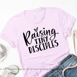 May include: A pink t-shirt with the text "Raising Tiny Disciples" in black lettering. The text is in a cursive font and includes a small heart above the word "Raising".