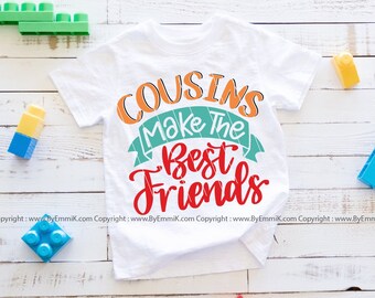 May include: White t-shirt with the text "COUSINS Make The Best Friends" in colorful lettering. The shirt is on a white wood background with colorful toy blocks.