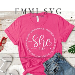 May include: A pink t-shirt with a white circle design that says "She is" surrounded by the words "Strong, Unstoppable, Powerful, Bold, Unique, Beautiful, Brilliant, Enough".
