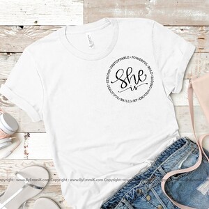 May include: White t-shirt with a black circle graphic that says "She is" surrounded by the words "Strong, Unstoppable, Powerful, Bold, Unique, Beautiful, Brilliant, Enough".