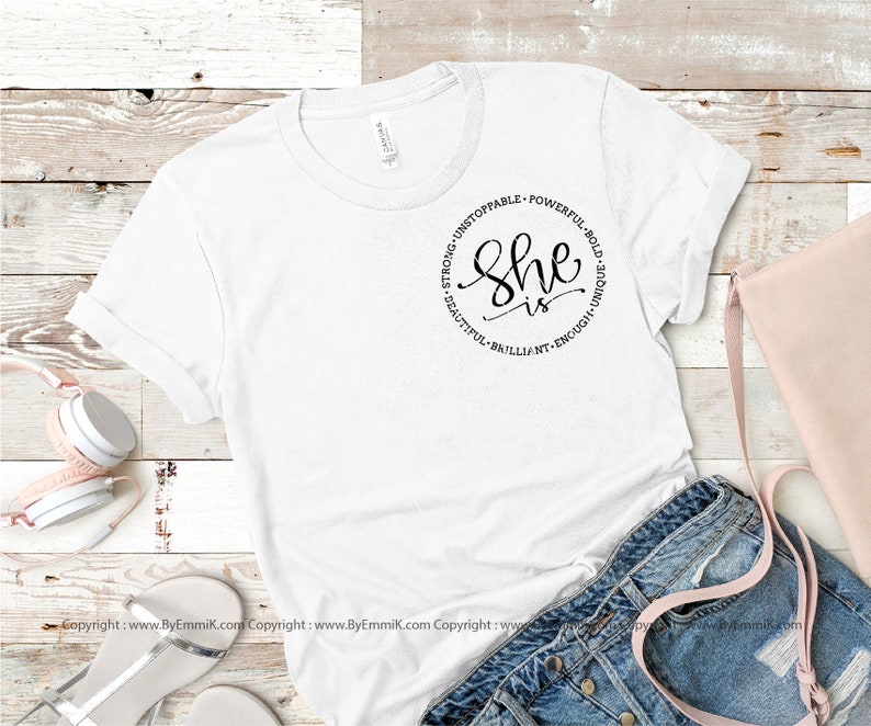 May include: White t-shirt with a black circle graphic that says "She is" surrounded by the words "Strong, Unstoppable, Powerful, Bold, Unique, Beautiful, Brilliant, Enough".
