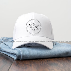 May include: White baseball cap with black text that reads "She is" surrounded by the words "Strong, Unstoppable, Powerful, Bold, Unique, Beautiful, Brilliant, Enough".