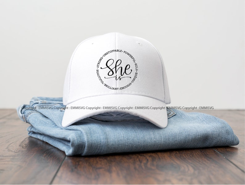 May include: White baseball cap with black text that reads "She is" surrounded by the words "Strong, Unstoppable, Powerful, Bold, Unique, Beautiful, Brilliant, Enough".