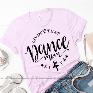 May include: A pink t-shirt with the text "Livin' That Dance Mom Life" in black lettering. A small silhouette of a dancer is in the center of the text.