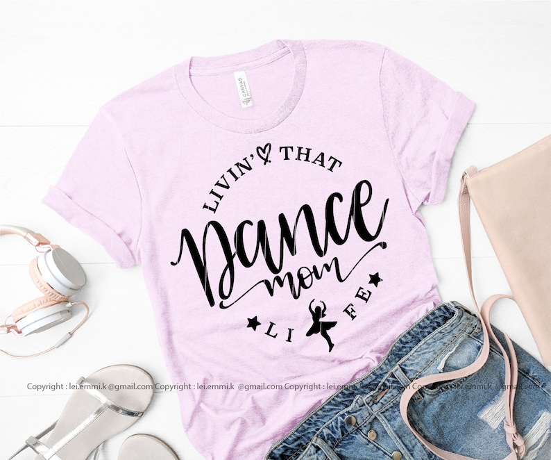 May include: A pink t-shirt with the text "Livin' That Dance Mom Life" in black lettering. A small silhouette of a dancer is in the center of the text.