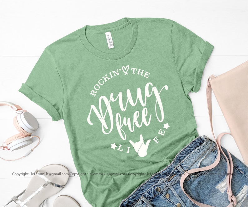 May include: A green t-shirt with the text "Rockin' the Drug Free Life" in white lettering. The text is in a circle with a heart and a star on either side. A white hand symbol is below the text.