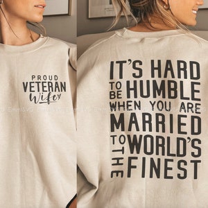 Pu&ograve; includere: Una felpa beige con testo nero. La parte anteriore della felpa dice "Proud Veteran Wife" e la parte posteriore dice "It's hard to be humble when you are married to the world's finest."